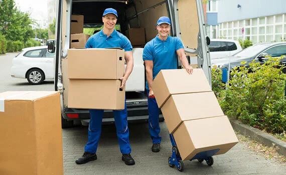 Two smiling movers in blue uniforms unloading boxes and a dolly from a van.
